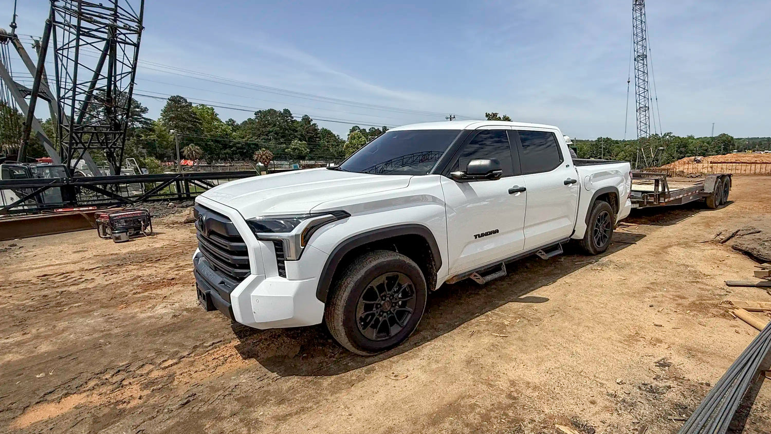 A white Toyota Tundra pickup truck, prior to having an ADAPT Tundra Ladder Rack installed, pulling a flatbed trailer with construction equipment in the background | Chandler Truck Accessories