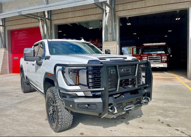 Photo of a white truck parked in front of a fire station. The white GMC truck has a bumper guard on the front that looks very rugged and durable. The truck is a GMC 2500/3500 (2024+) with a Front Bumper - Full Guard Bumper made by Chandler Truck Accessories.