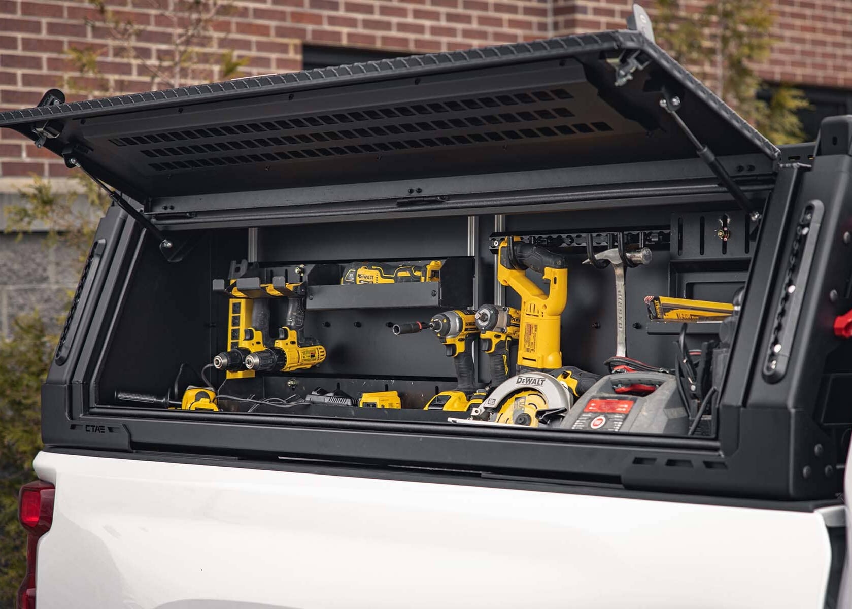 An open tool box that is mounted onto the side of a truck, the truck is outside and parked next to a brick building. The truck tool box is open and features many tools. 