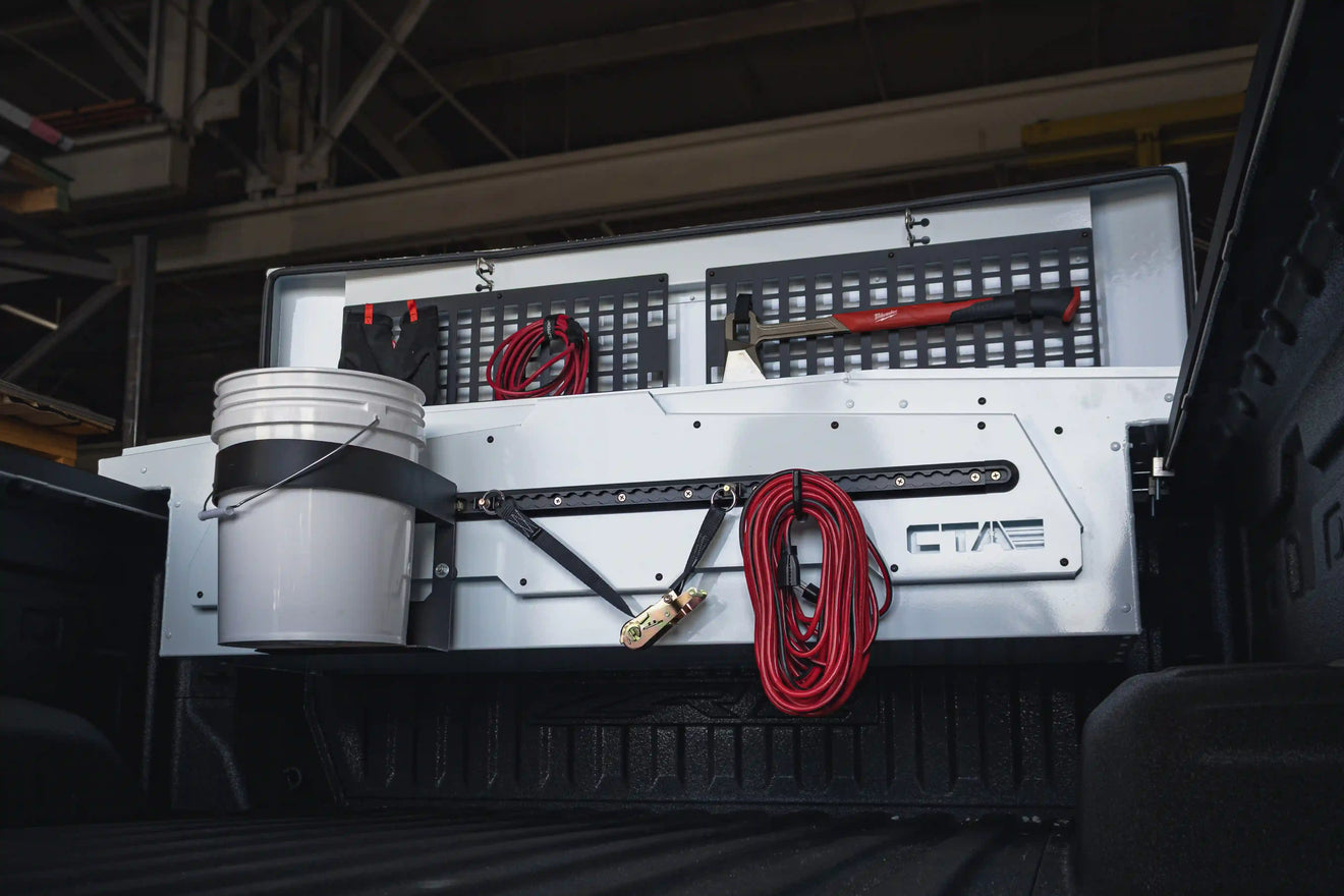 White ADAPT truck bed tool box in pickup truck with external mounted bucket holder, ratchet strap, and extension cord. A milwaukee axe is mounted on the lid along with gloves and another extension cord.